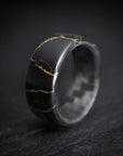 A black and gold ring with a carbon fiber core and rose gold colored accents, displayed on a dark background.